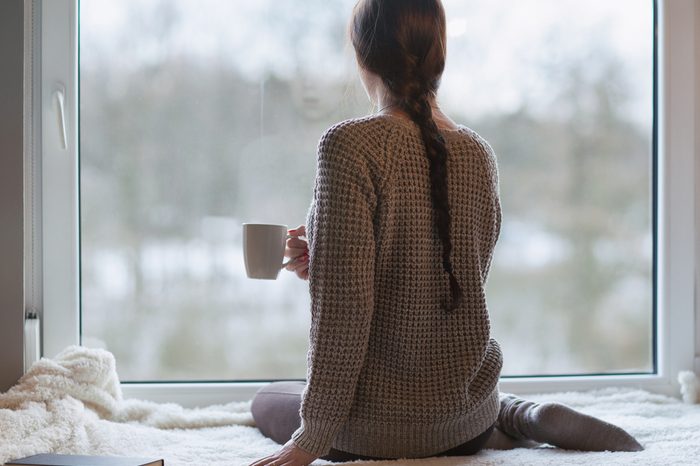 Thoughtful young brunette woman with book and cup of coffee looking through the window, blurry winter forrest landscape outside