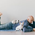 Adult bald man in fashionable clothing lying on yoga mat. Young guy reading book. Two white male chihuahua puppies at home. Pet sleeping with owner. Little lovely furry dog sitting on wooden floor