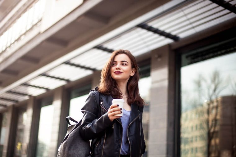 young beautiful brunette drinking coffee walking around the city.leather jacket,urban backpack , bright red lips Posing against the window of the boutique Model looking aside
