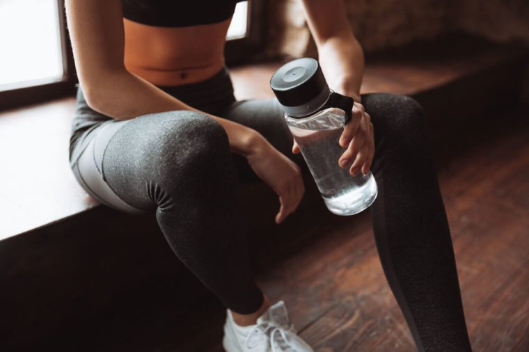 Cropped photo of attractive fitness woman sitting in gym and holding bottle of water.