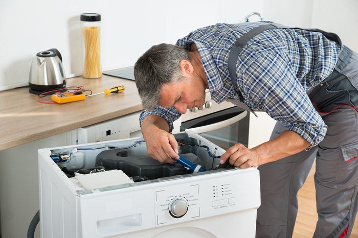 Full length of handyman checking washing machine with flashlight in kitchen