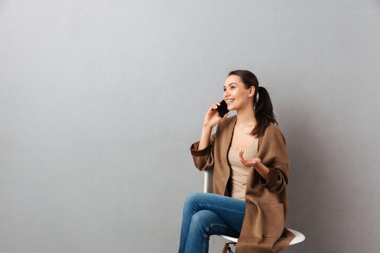 Portrait of a smiling casual asian woman talking on mobile phone while sitting on a chair over gray background