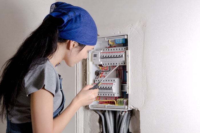 Young woman holding a screwdriver looking at an open electrical box and checking the circuit breakers