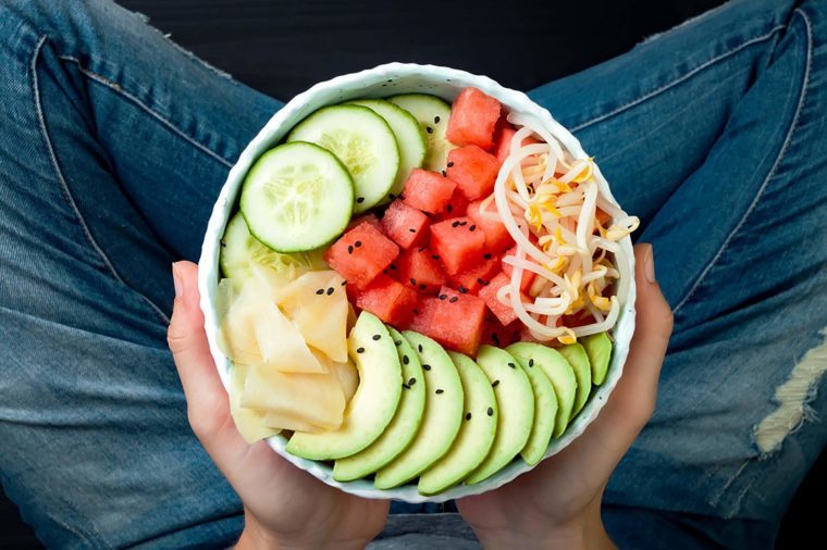 Girl in jeans holding hawaiian watermelon poke bowl with avocado, cucumber, mung bean sprouts and pickled ginger. Top view, overhead