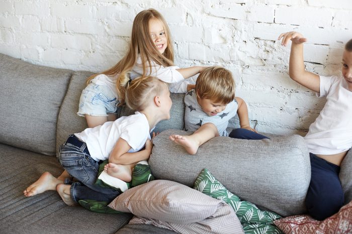 Action shot of four naughty children siblings fooling around at home, turning living room upside down. Restless cheerful kids in casual clothes playing, having fun, fighting with pillows on sofa