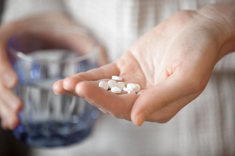 Healthcare, treatment, supplements concept photo. Woman arm holding heap of small round meds and glass of water before taking medication, shallow depth of field, focus on medicine