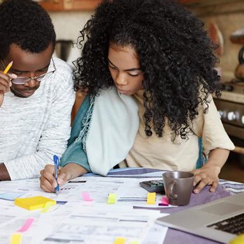 Young confident African housewife with Afro hairstyle helping her husband to manage domestic finances, calculating and making notes with pen, both sitting at kitchen table with laptop and papers