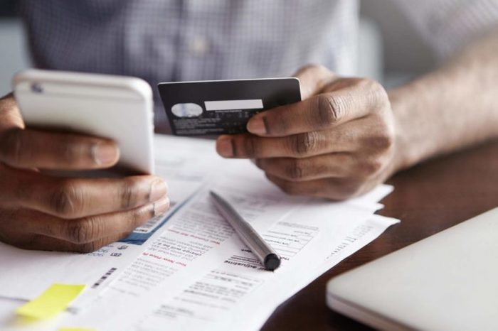Online payment and shopping concept. Cropped shot of African-American male holding cell phone in one hand and credit card in other, making transaction, suing mobile banking app during lunch at cafe