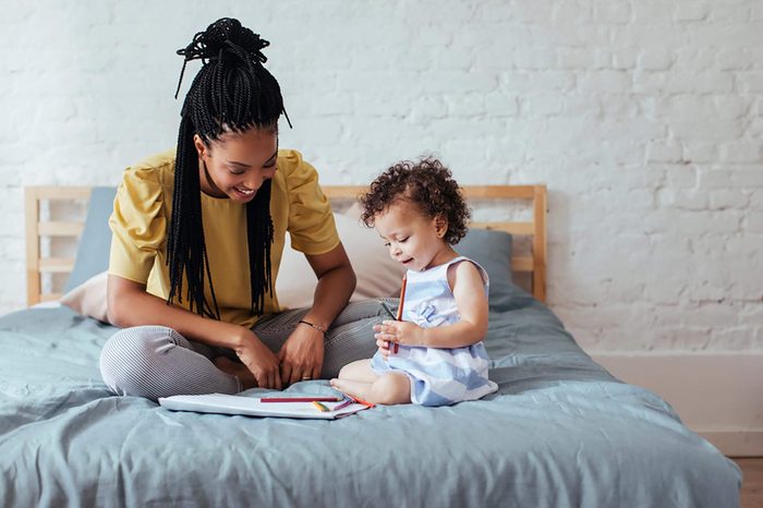 Beautiful African woman sitting on bed with her baby daughter and drawing on paper.