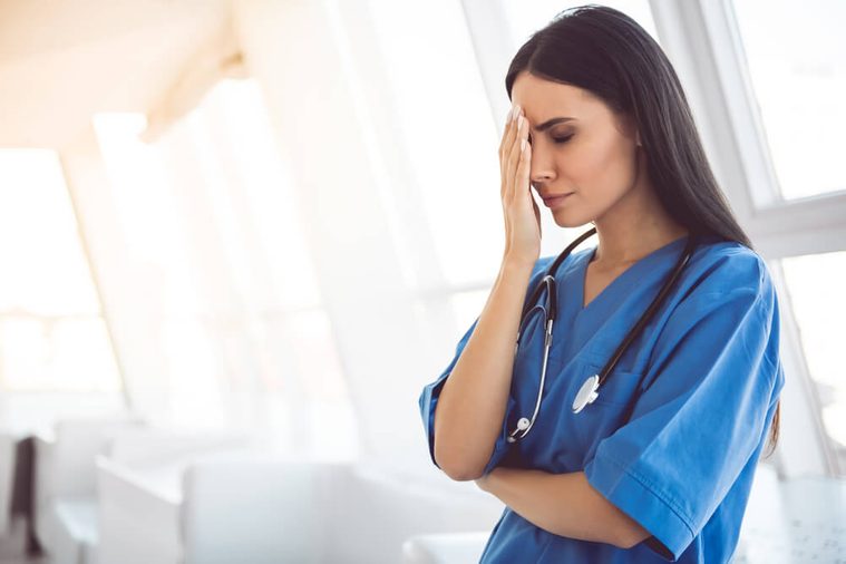 Beautiful doctor in blue scrubs is covering her face while standing in hospital corridor