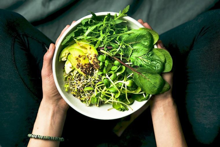Green vegan breakfast meal in bowl with spinach, arugula, avocado, seeds and sprouts. Girl in leggins holding plate with hands visible, top view. Clean eating, dieting, vegan food concept