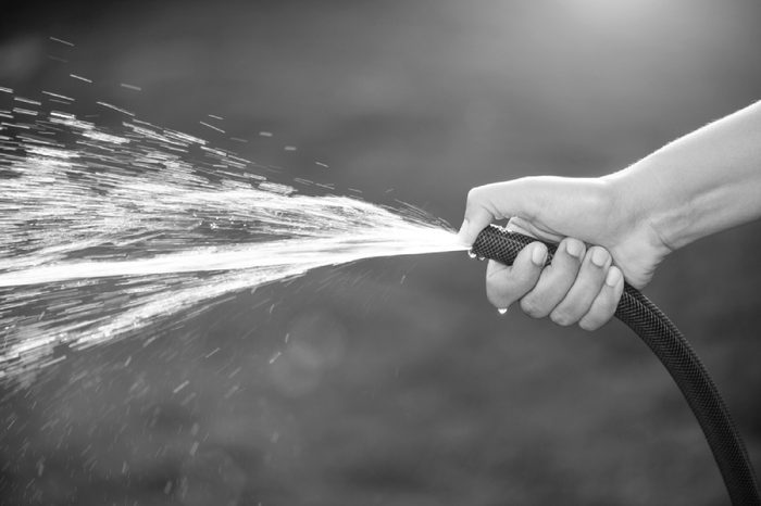 Woman hand holding rubber water hose and using finger close end of rubber water hose to make water spray with sunlight and green grass field in background. World Water Day conpect.