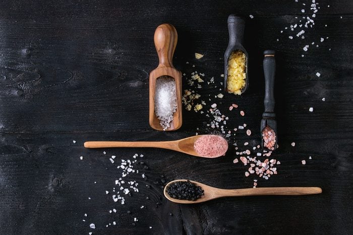 Variety of different colorful salt yellow saffron, pink, black himalayan, white sea and fleur de sel in wooden spoons over black burned wood background. Top view with space
