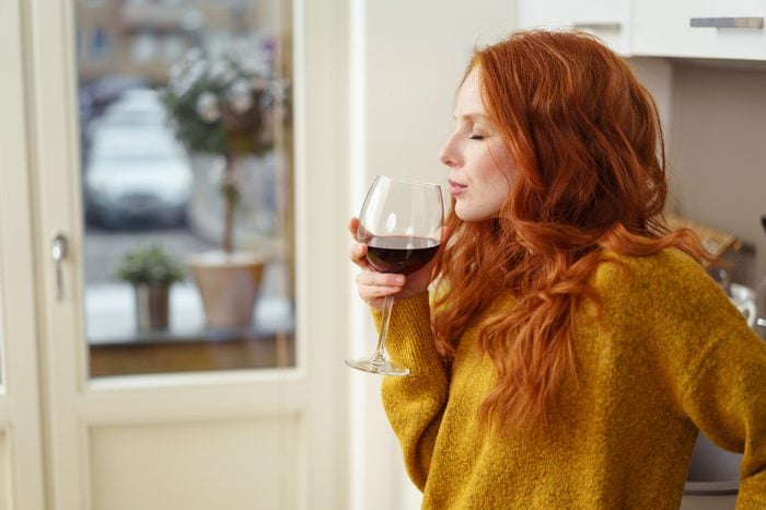 Young redhead woman standing in her apartment sipping a glass of red wine with her eyes closed in pleasure, side view