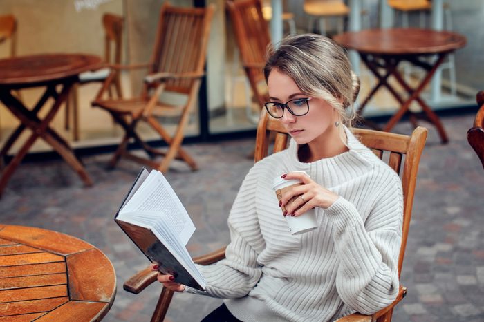 Beautiful young woman reading a book with a cup of coffee sitting in the street cafe