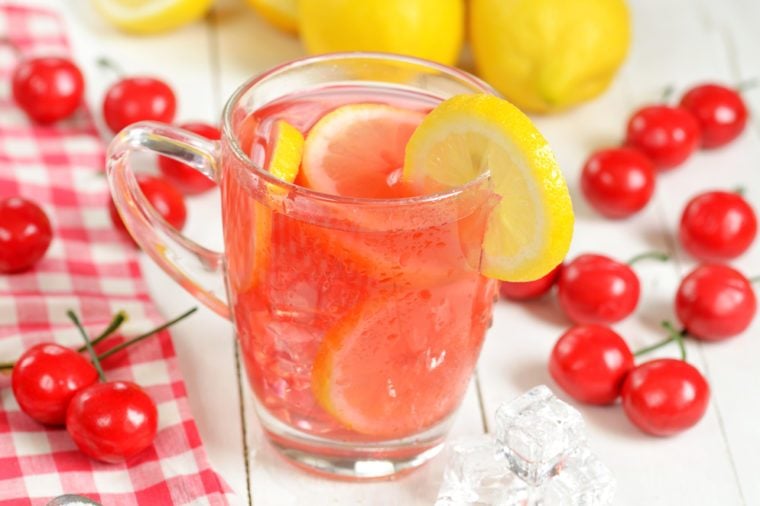 Fruit cherry ice tea with slice of lemon in glass mug, red checkered tablecloth, fresh cherries and lemons in foreground
