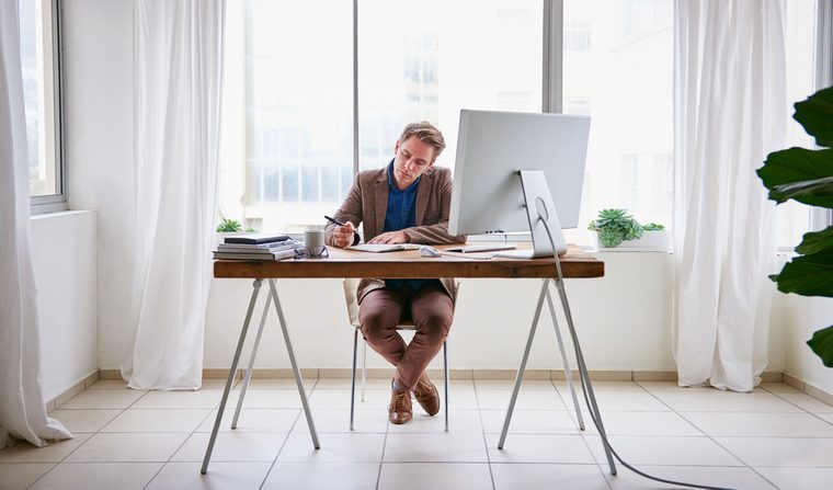 Full length image of a young male entrepreneur sitting at his desk in a contemporary work space for one person and writing in his journal