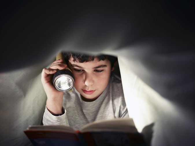 caucasian child reading book under the covers at night. Front view, copy space