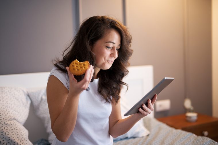 Pretty satisfied middle aged women sitting on the bed and looking on a table before sleeping while eating a biscuit.