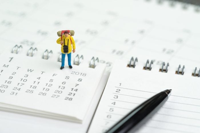 Travel, vacation or holiday calendar year plan concept, miniature traveller backpacker man figure standing on pile of calendars and numbers listed of wish list to go and pen to write destinations.