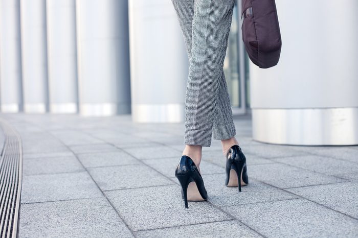 Close up of female legs of worker standing near her office. The woman is wearing formalwear and shoes on high heels. She is holding a handbag. Copy space in left side