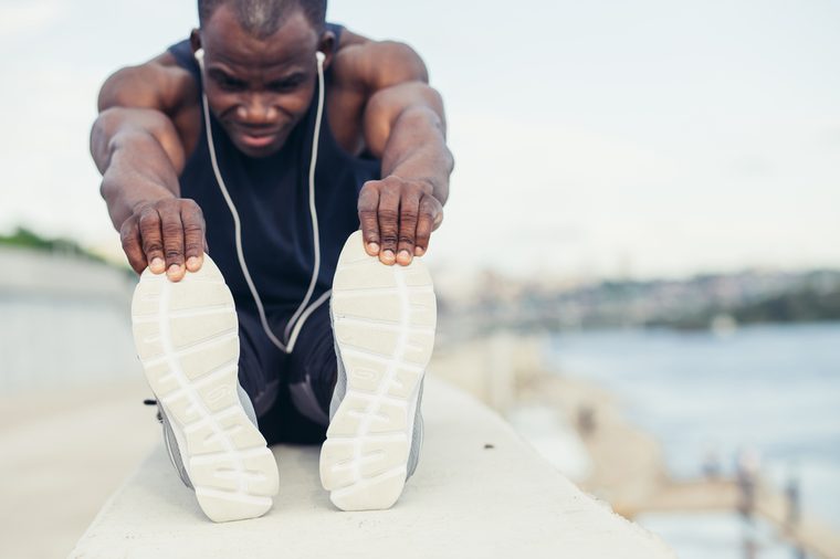 Black man doing stretching before running in urban background. Young male exercising listening to music with headphones.