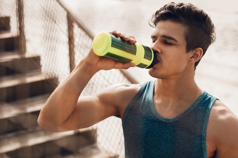 Close up shot of fit young man drinking water by the beach after workout. Runner drinking water after exercising.