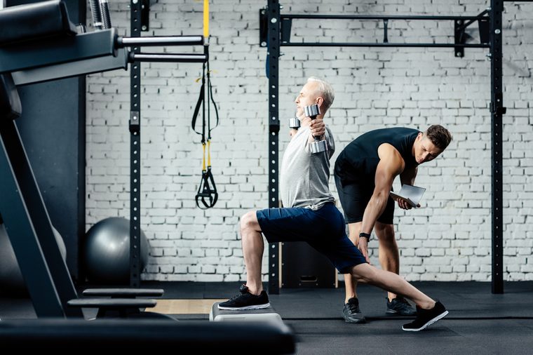 senior sportsman doing lunges with dumbbells in sport center with trainer near