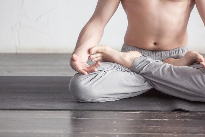 A young strong man doing yoga exercises - lotus pose and meditates with padmasana legs. Studio full length shot over white brick background and black floor, close up.