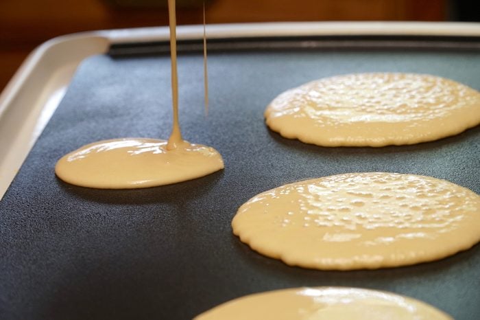 Pancake batter baking mix being poured from a bowl onto a hot electric griddle cooking delicious breakfast meal for a family.