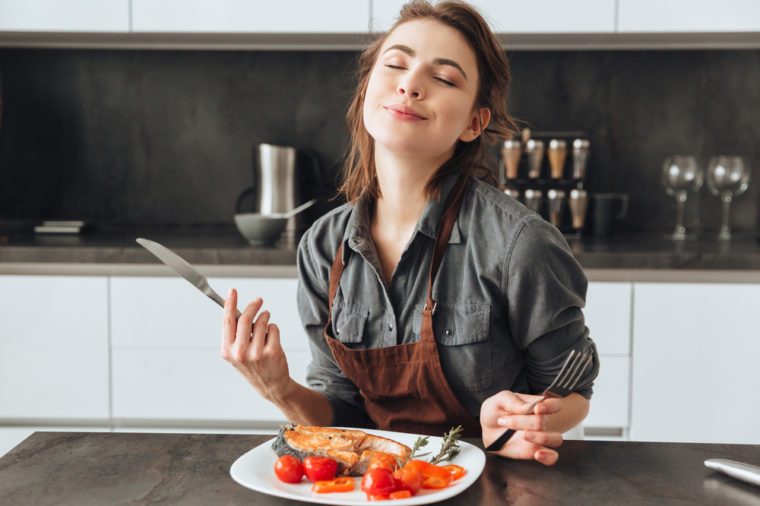 Image of pretty young woman sitting in kitchen while eating fish and tomatoes.