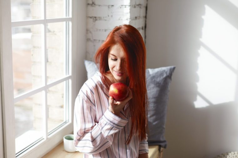 Picture of gorgeous smiling young redhead female wearing stylish pajamas posing by large window in her room, holding big ripe red apple, having excited anticipated look, enjoying tasty healthy fruit
