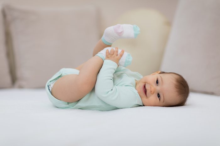 Happy baby lying on white sheet and holding her legs / Playful baby lying down in bed