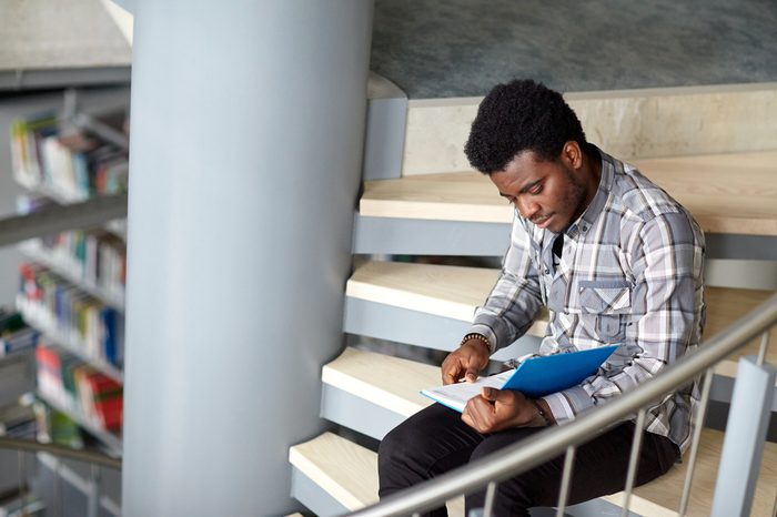 education, high school, university, learning and people concept - happy african american student boy or young man reading book sitting on stairs at library