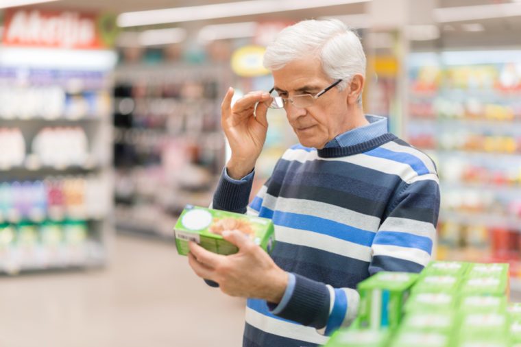 Elderly man shopping in local supermarket. He is holding box and reading nutrition label.