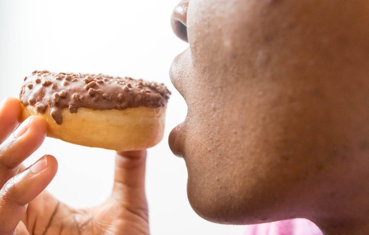 Close up picture of a African American woman eating holding a delicious doughnut with chocolate and biting processed foods weight loss diet concept