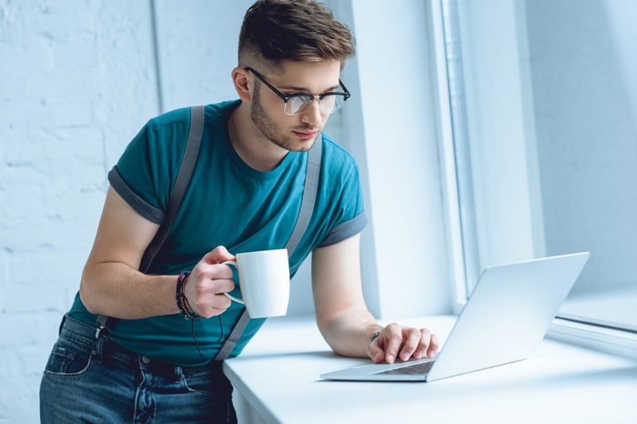 young man in eyeglasses holding cup of coffee and using laptop on windowsill