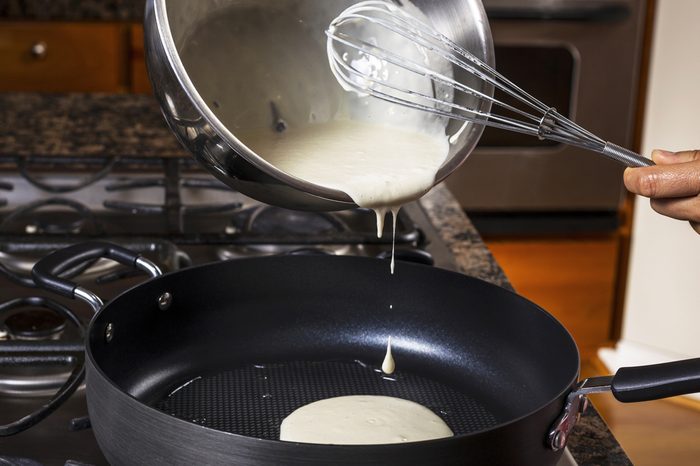 Pouring pancake batter into frying pan with stove top in background