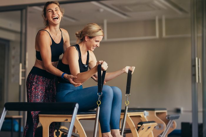 Laughing female trainer helping a woman in pulling stretch bands sitting on pilates training machine. Smiling woman at the gym doing pilates training with her trainer.