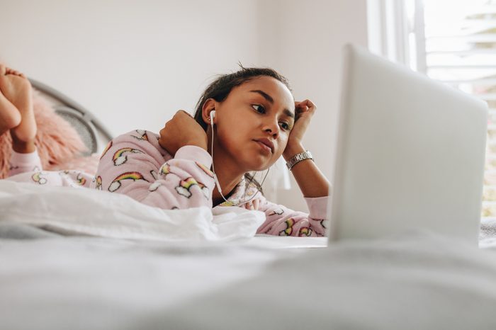 Young girl in bed watching a movie on laptop. Girl using laptop lying on bed while listening to music using earphones.