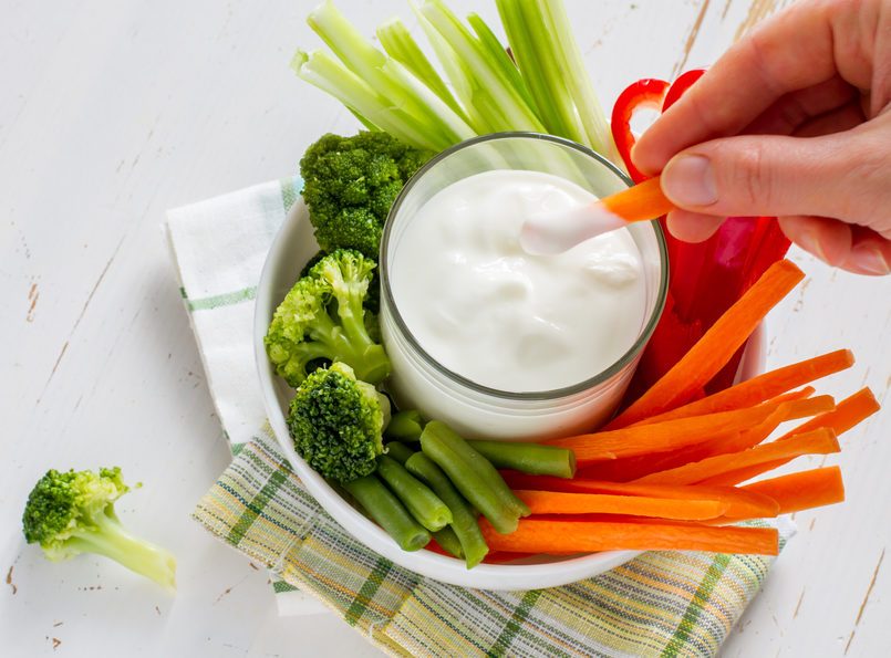 Hand holding carrot, white bowl with carrot, celery, pepper, broccoli and green beans, yogurt sauce, white wood background