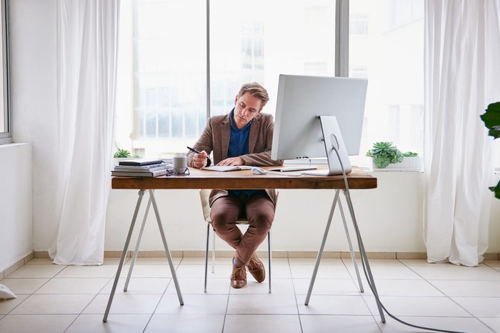 Full length image of a young male entrepreneur sitting at his desk in a contemporary work space for one person and writing in his journal
