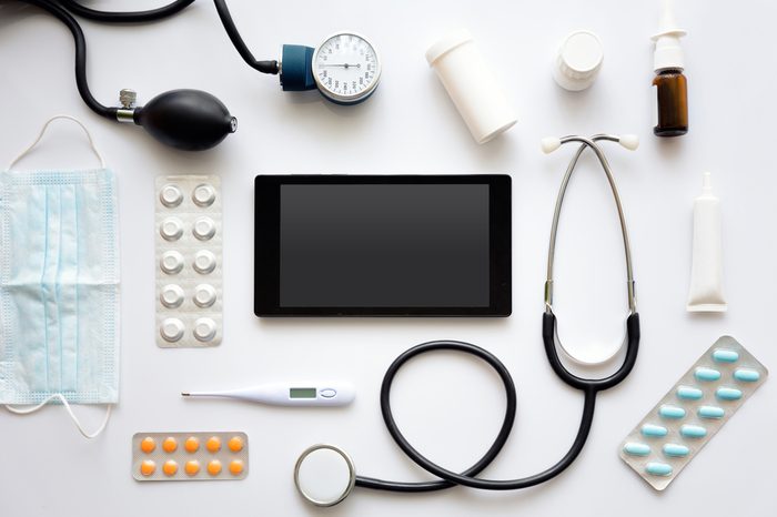 E-medicine concept.Black tablet mock up,together with stethoscope,thermometer,various medications and surgical mask around it,laid flat on top of white surface.