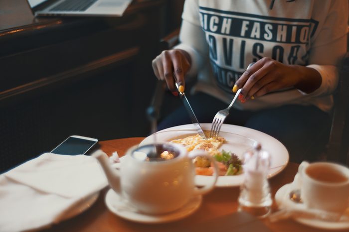 Hands of young black girl in a cafe at daytime, wearing a long sleeve shirt and jeans. She is eating omelet with vegetables using knife and fork and drinking tea, nail art, vintage colour