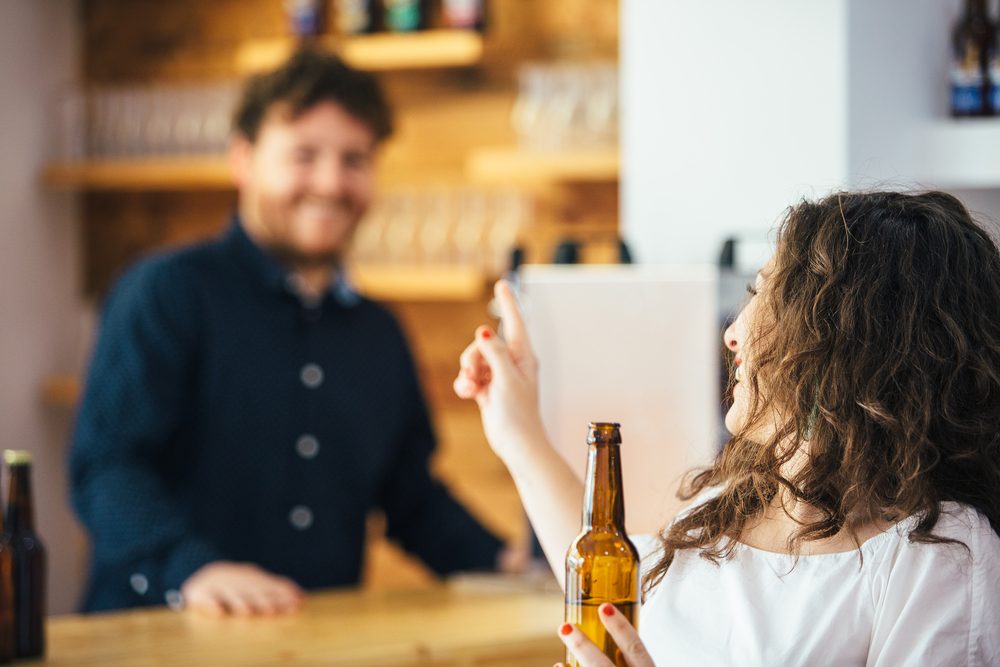 Curly-haired brunette with bottled beer talking and smiling at unfocused bartender