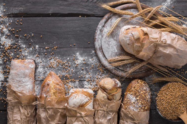 Bread background. Brown and white whole grain loaves wrapped in kraft paper composition on rustic dark wood with wheat ears scattered around. Baking and home bread making concept. Soft toning