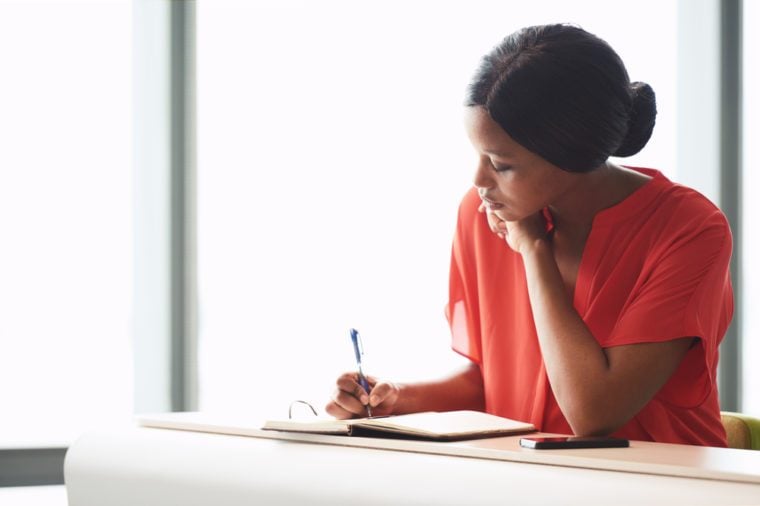 Self employed black female entrepreneur busy writing in her notebook while seated at a desk with a large bright window behind her in the background.