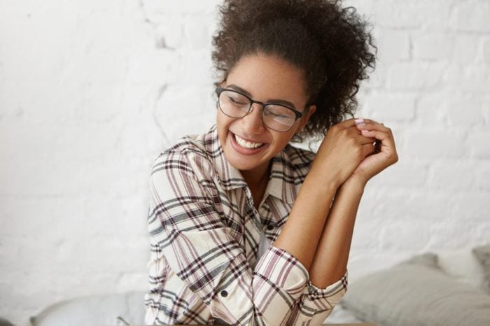 Positive human facial expressions and emotions. Indoor shot of cute charming Afro-American girl wearing stylish plaid shirt and eyeglasses, closing eyes in pleasure and excitement, smiling cheerfully