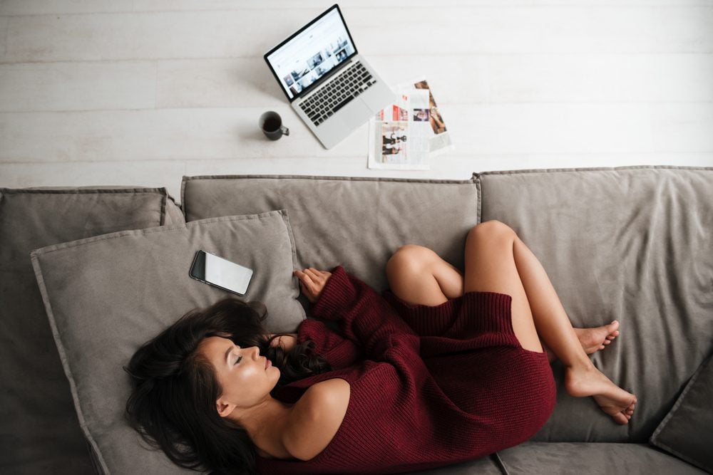 Top view of a beautiful asian woman in sweater sleeping on a couch at home with blank screen mobile phone and laptop