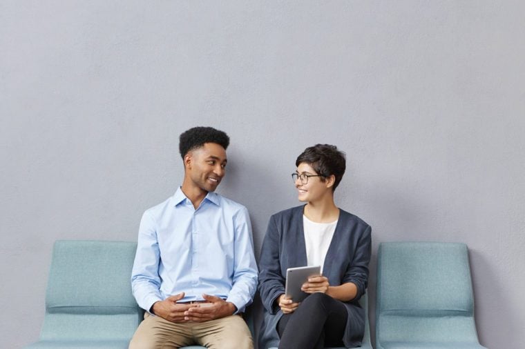 Candid shot of positive young African man and Caucasian woman job candidates wearing formal clothes sitting in queue on chairs in waiting room for a long time, talking and amusing themselves